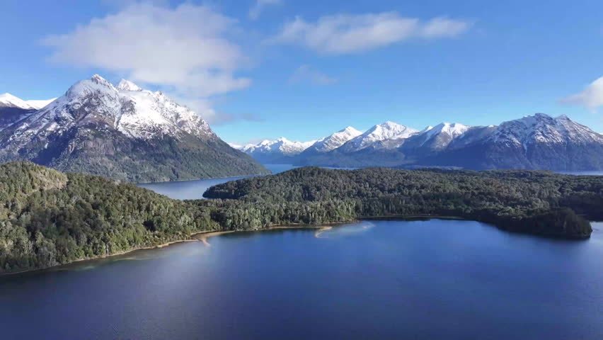 An aerial perspective showcases majestic snow-capped mountains surrounding a tranquil lake, with lush greenery along the shoreline under a clear blue sky.