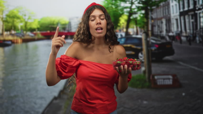 Woman holding strawberries with bare shoulder exposed and hand extended toward camera on street canal; confidence summer invitation.