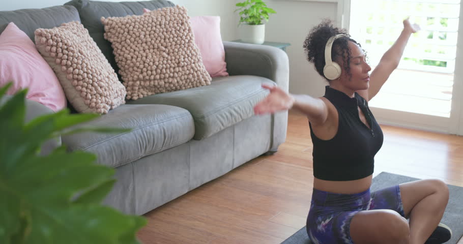African American woman breathing stretching, meditating for calm on mat with headset, copy space. Yoga, mindfulness, zen, wellness, serenity, home, sunlit