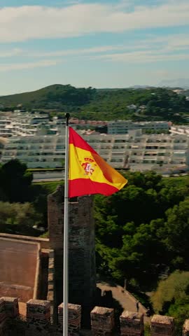 Vertical aerial orbit of Spanish flag waving above historic fortress against the Mediterranean coastline, Sohail Castle, Fuengirola, Andalusia, Costa del Sol, Spain