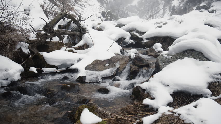 Cold mountain stream water flowing rapidly over rocks in a pristine winter landscape. The creek banks are covered with fresh snow and ice, creating a serene and wild natural scene