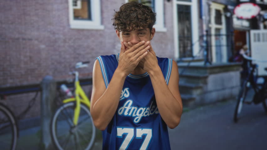 Teenage boy in blue basketball jersey with hands covering mouth on a street with bicycles and building steps visible; surprise youthful energy.