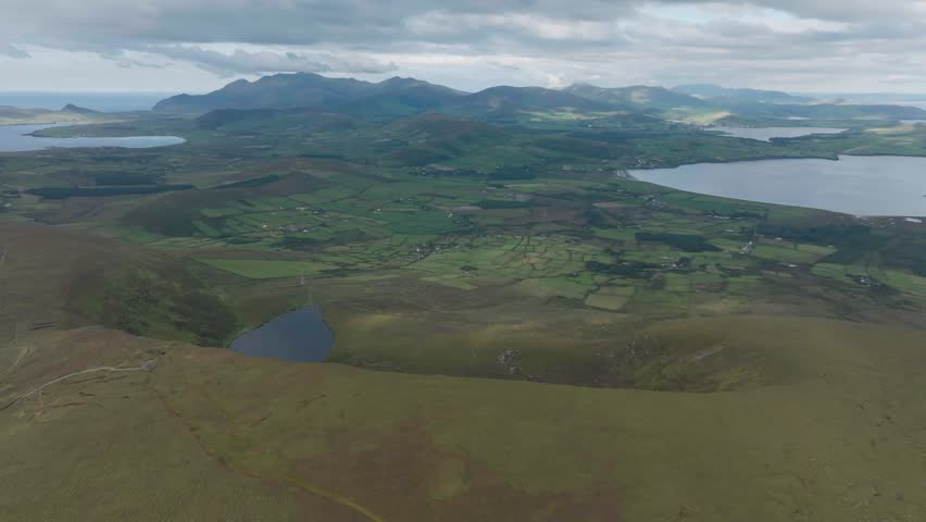Breathtaking aerial view of the Dingle Peninsula in Ireland featuring lush green fields, rolling mountains, and serene lakes under a moody sky. Perfect for travel documentaries and nature content.