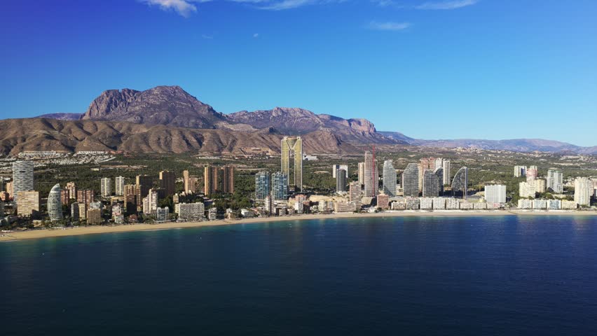 Aerial view of the Benidorm cityscape on Costa Blanca Spain featuring beachfront buildings and a mountain backdrop.The Mediterranean Sea under a clear blue sky on a sunny day