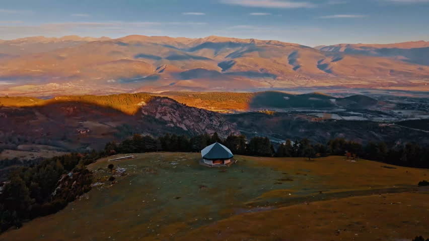 A beautiful aerial view of a mountain landscape featuring a yurt surrounded by lush greenery and distant peaks illuminated by the warm glow of sunset.