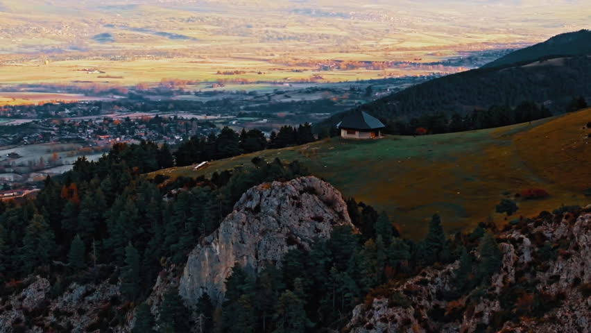 Aerial view of a serene mountain landscape featuring a small cabin surrounded by trees and rolling hills. The scene captures the tranquility of nature.