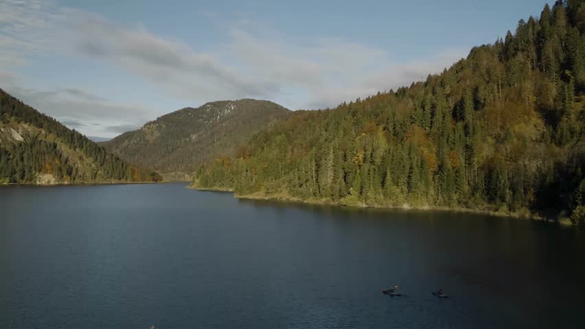 Kayakers glide across a vast, deep blue lake bordered by a dense forest displaying vibrant autumn foliage and majestic, cloud-kissed mountains