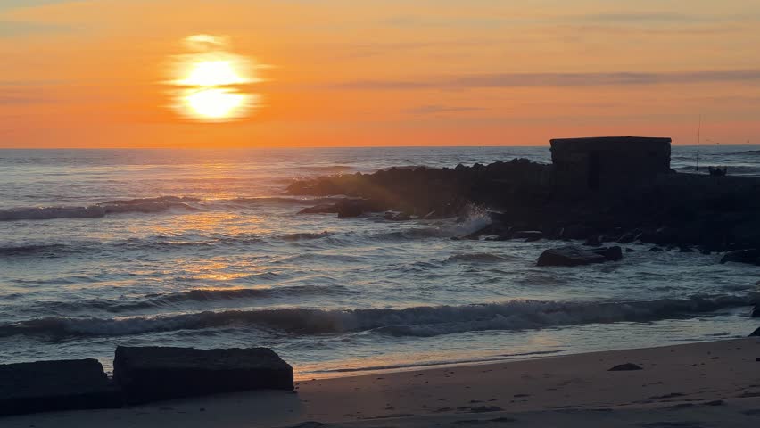 Static shot of sunset over ocean waves breaking against stone breakwater on sandy beach in Barbate, Cadiz, Spain.