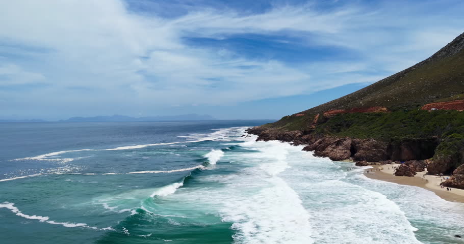 Aerial drone view of turquoise waves rolling onto a sandy cove and rocky shoreline at Kogel Bay along Clarence Drive in the Western Cape, South Africa. Dramatic coastal cliffs, clear blue ocean, and scattered beachgoers create a scenic summer seascape ideal for travel and tourism concepts