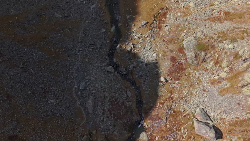 Top-down drone view following mountain stream near Col du Glandon in the Belledonne range, French Alps. Aerial alpine landscape with rocky terrain and autumn tones.