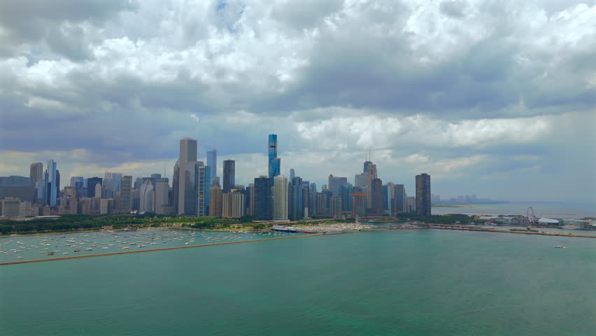 Drone aerial view of Chicago Illinois skyline stretching along Lake Michigan shoreline, turquoise water, dramatic cloudy sky and wide urban panorama in daylight.