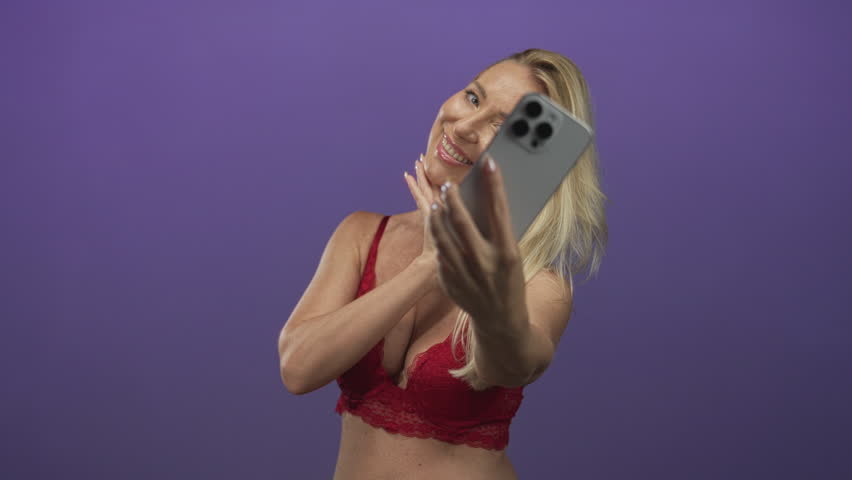 Blonde woman taking selfie with smartphone showing red lingerie cleavage in studio; playful confidence.