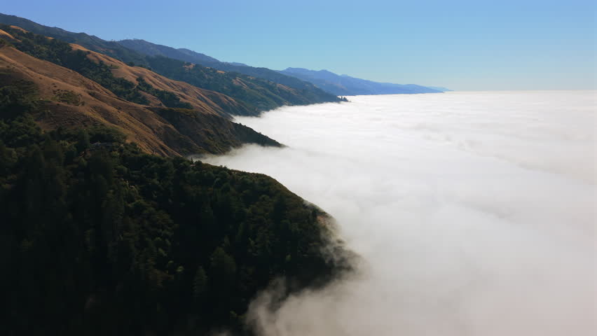 Aerial view of Big Sur California coastline rising above dense sea of fog. Golden hills meet Pacific horizon while Highway 1 traces dramatic coastal mountains.