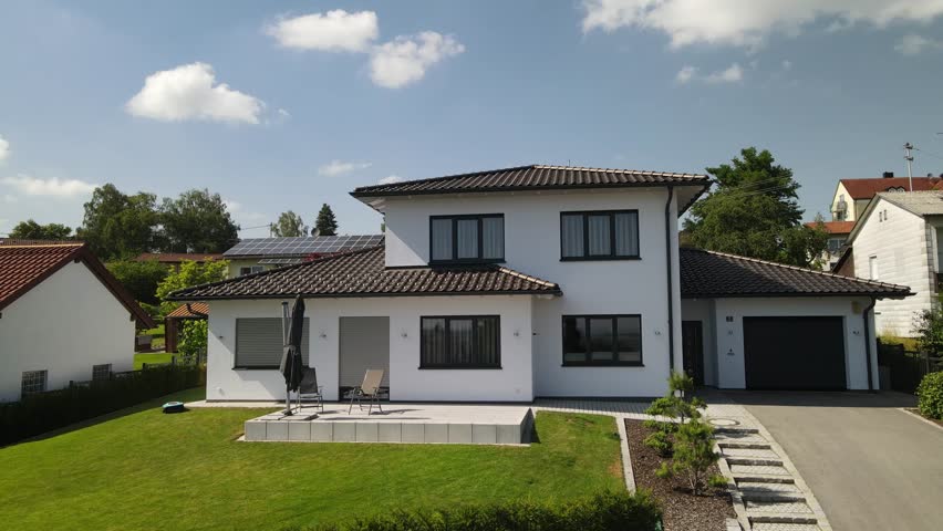 Modern two story detached house in a quiet German village suburb with landscaped garden, driveway and garage under blue summer sky.
