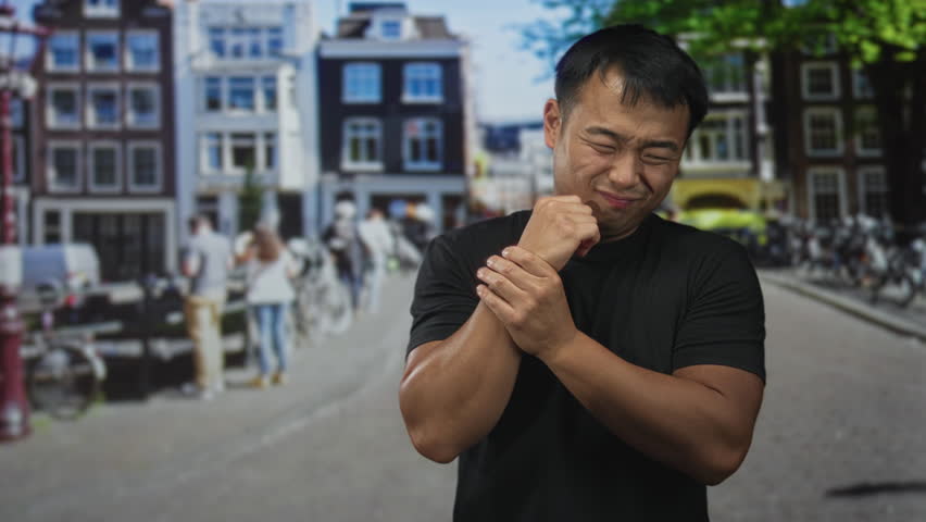 Young chinese man in black shirt grasps wrist on sunlit urban street by canal buildings; discomfort.