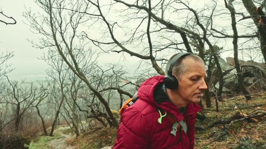 Focused senior man with headphones enjoying music while exploring misty hills during a rainy day for motivation and inner peace.