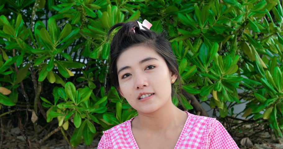 Young asian woman smiling outdoors by lush foliage wearing pink gingham dress and hair clip, candid headandshoulders portrait against tropical shrub backdrop, soft natural light, relaxed friendly