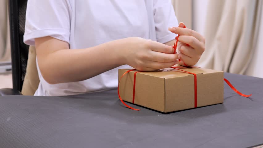 Person is tying a red ribbon around a brown box. The box is on a table. The person is wearing a white shirt