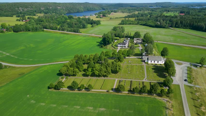 Gunnarsnäs Church, Mellerud, Sweden — aerial parallax, white church, tree-lined cemetery, vast green agricultural fields, lakes and forest horizon visible, sunny summer day