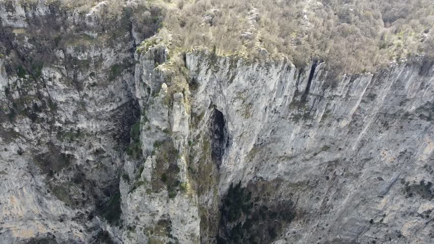 A cinematic drone close-up of the towering limestone walls of Vikos Gorge. Highlighting the jagged rock textures and the sheer verticality of the world