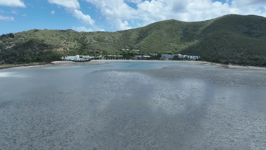 Slow drone push-in in daytime over tidal reef flats toward Hayman Island resort, revealing the shallow lagoon bay and beachfront backed by green hills, Whitsundays, Queensland, Australia.