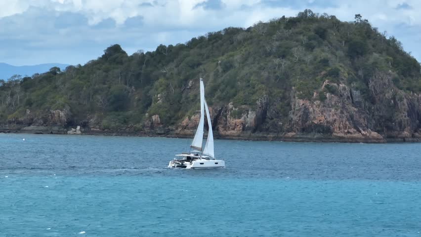 Telephoto drone shot in daytime with a slow drift, holding on a white sailboat cruising across turquoise water with a rugged green island backdrop, Whitsundays, Queensland, Australia.
