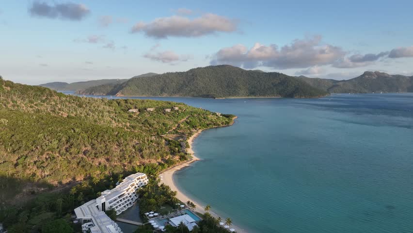 Slow drone pullback while orbiting right, revealing Hayman Island’s tropical coastline, sandy beach and luxury resort beside calm turquoise ocean waters in the Whitsundays, Queensland, Australia.