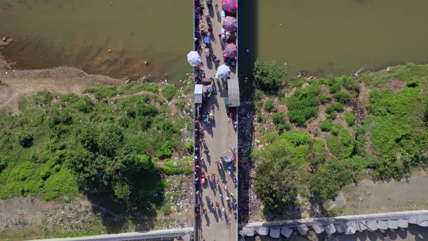 Top-down aerial view of Dominican–Haitian border in Dajabón. Crowds gather along narrow bridge over river as vendors and traders prepare for busy cross-border market day under bright sun.