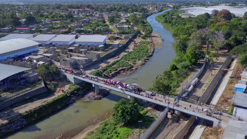 Aerial view of Dominican–Haitian border in Dajabón as crowds gather on bridge for market day. Traders, vehicles and colorful umbrellas fill crossing during preparations for cross-border commerce.