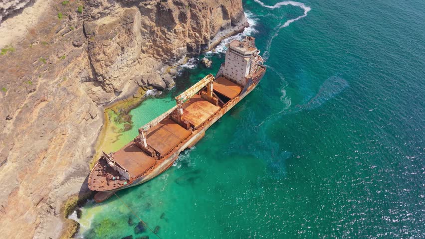 Aerial view of the rusted MV Jernas cargo ship abandoned against the rocky cliffs along the turquoise coastline of Salalah, Oman.