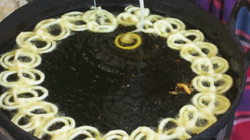Indian halwai preparing crispy jalebi in hot oil at a roadside sweet shop, shaping spiral batter by hand in a large kadai during busy market hours.