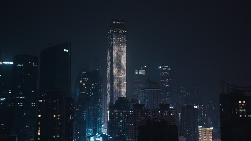 Aerial drone night shot of Chongqing World Financial Center skyscraper with illuminated CHONGQING signage on facade amid surrounding skyscrapers in Jiefangbei district