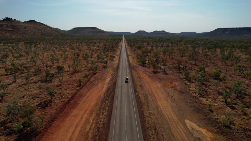 Van drives along empty Victoria Highway in Northern Territory, surrounded by vast bushland, red dirt and distant mountain ranges. Captured by drone.