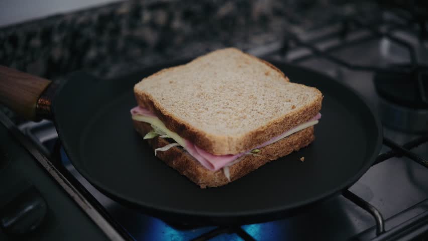 Close up of a person cooking a grilled ham and cheese sandwich in a frying pan. They add butter, flip it with a spatula, and press it down to toast.