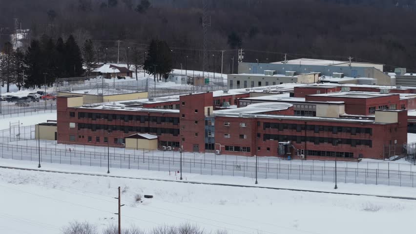 Aerial view of brick Federal Correctional Institution complex in Loretto, Pennsylvania, surrounded by security fencing and snow-covered grounds during winter, with wooded hills in background.Wide shot