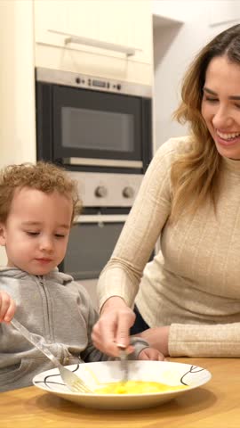 Adorable little boy learning to cook with his mother, happily whisking eggs in a bowl on the kitchen counter while preparing a family meal and sharing a moment of bonding