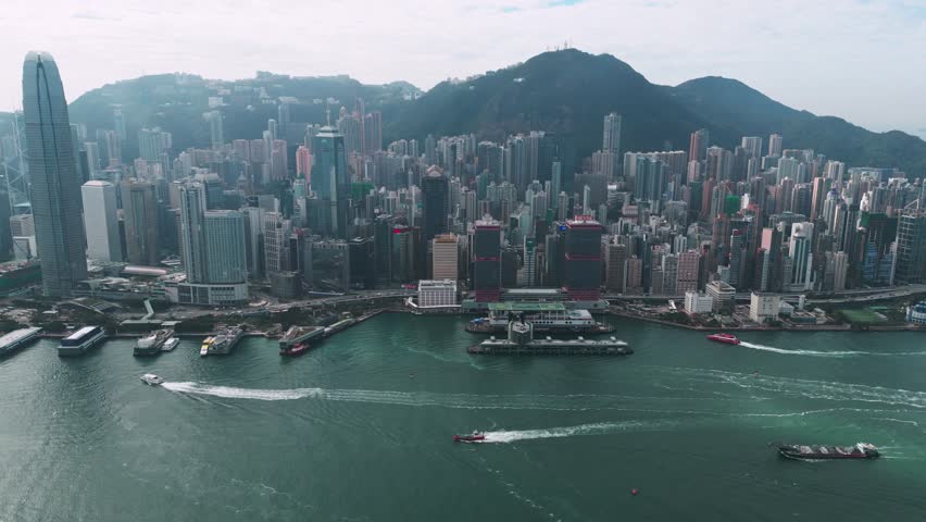 Panoramic aerial view of the skyscrapers and Victoria Harbour in downtown Hong Kong on a sunny day, showcasing the iconic skyline and waterfront.