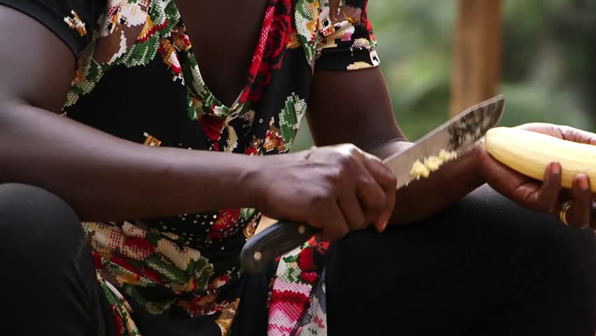 Close-up of Ghanaian woman peeling cassava with a machete knife, traditional food preparation outdoors in rural West Africa.