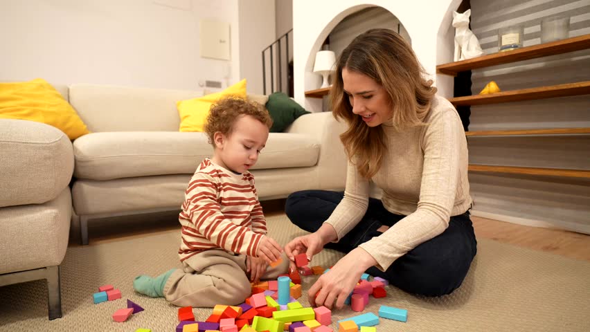 Happy mother and toddler son sit on the living room floor stacking colorful wooden blocks, sharing playful bonding time that encourages learning, development and joy
