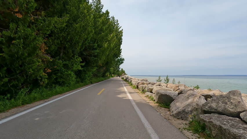 POV Biking on M-185 Highway Along Lake Huron Coast, Mackinac Island, Michigan, USA. A slow-motion, point-of-view shot from a bicycle riding along the shoreline of M-185 on Mackinac Island, Michigan. The paved road stretches ahead with several cyclists and pedestrians visible in the distance, flanked by a rocky coastline and green forest.