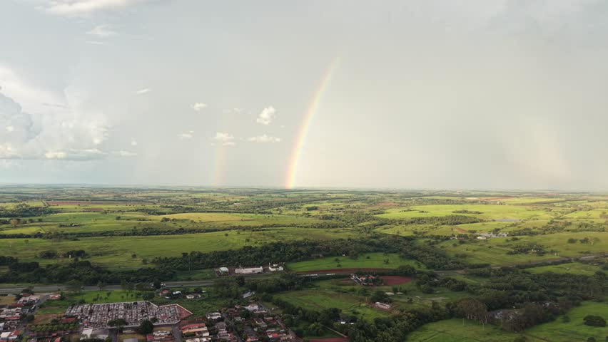This video captures a breathtaking rainbow arcing over a picturesque landscape with a small town nestled in the valley below. The scene showcases a serene and peaceful atmosphere with lush green fields, trees, and homes.