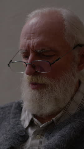 Vertical tilt down slowmo shot of concentrated grey-haired Caucasian man in eyeglasses and knitwear sitting in dimly lit room and looking at documents, analyzing monthly expenses