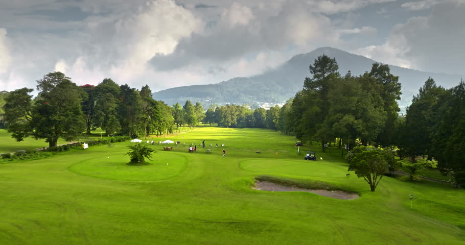 Aerial drone flight over Bali golf courses. People playing golf swings and relaxing under umbrellas at a lush green driving range with dense trees and a dramatic mountain landscape under cloudy sky