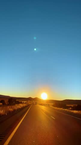 Sunset over highway leading through desert plains and mountains. Wide perspective of a multilane road heading directly towards the sun on the horizon in a desert landscape.