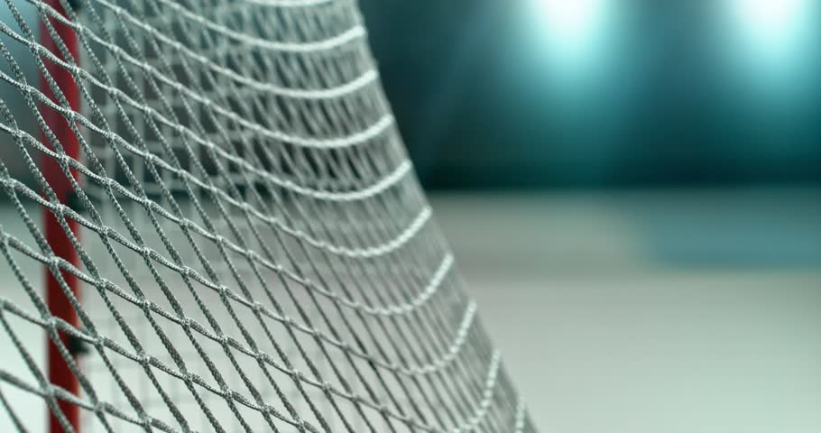 Extreme super slow motion close-up of an ice hockey puck striking the goal net at 1000 fps, with ice particles exploding on impact and a blurred winter stadium and rink in the background.