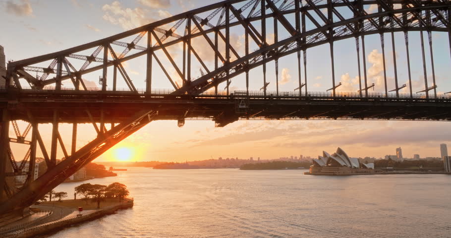 Train crossing Sydney Harbour Bridge spanning across the water, iconic Opera House and city skyline in the background, evening cityscape illuminated by the warm light of a golden sunset. City panorama