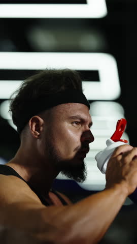 Focused athlete in a dim gym drinks from a protein shaker, then turns in profile with intense concentration, highlighting muscular strength, sweat, and determination under bright strip lights.