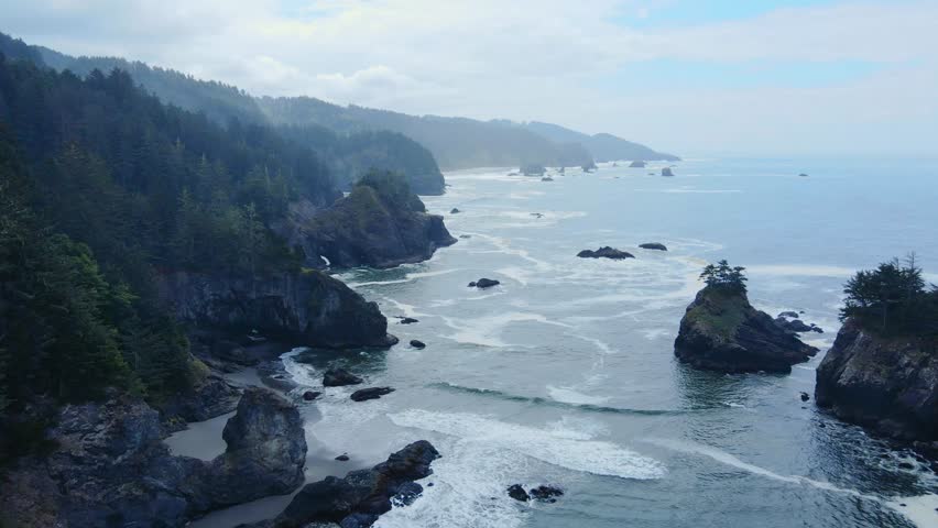 Aerial view of rugged pacific coastline with forested cliffs, sea stacks, and rolling surf under hazy sky, Oregon, USA