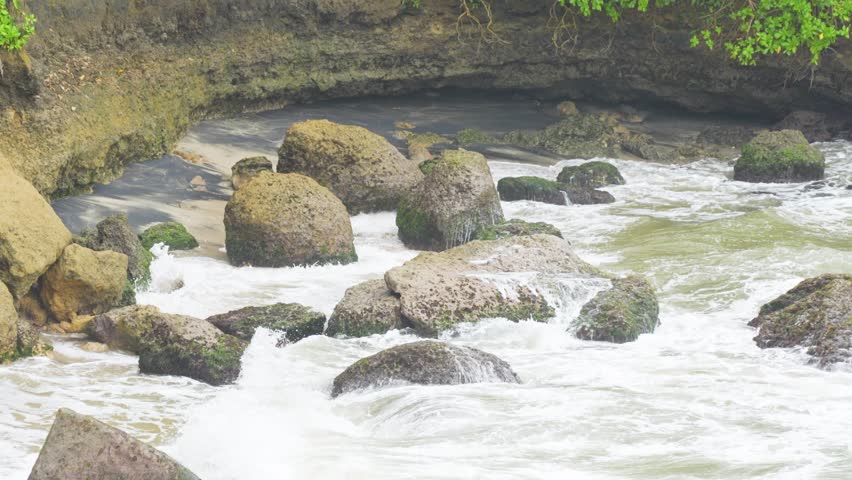 Ocean waves crashing against mossy rocks on a sandy beach below a weathered cliff. Natural coastal shoreline with dramatic rock formations.