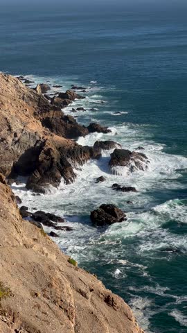 Rugged rocky coastline with steep cliffs and boulders as ocean waves crash and create white surf, viewed from above on a clear day.